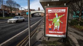 A bus shelter screen of the artwork ‘GROSSLY UNDERVALUED DOMESTIC PRODUCT’ (2022) by Bobby Baker in Manchester. A colourful sketched character on a red background. The central figure is adorned with cooking utensils, washing and other domestic symbols. She has several arms; one holds a bulging shopping basket and another arm leans on a walking cane. A child's hand tightly grasps onto another hand and one arm holds a large flag with text across it. The text reads: 'GROSSLY UNDERVALUED DOMESTIC PRODUCT'.